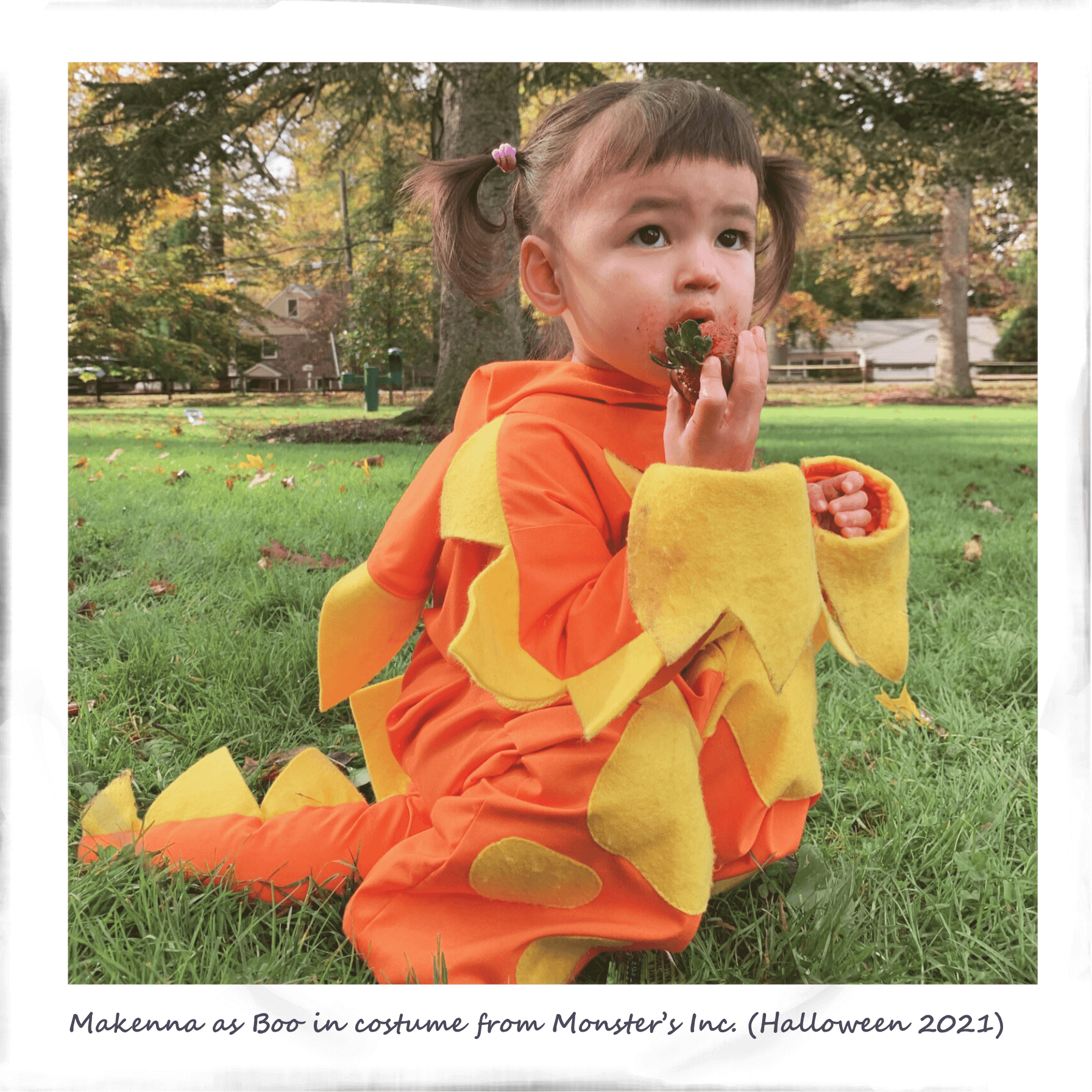 Little girl in a cute orange dinosaur costume sitting on grass and eating strawberry, in a park during autumn